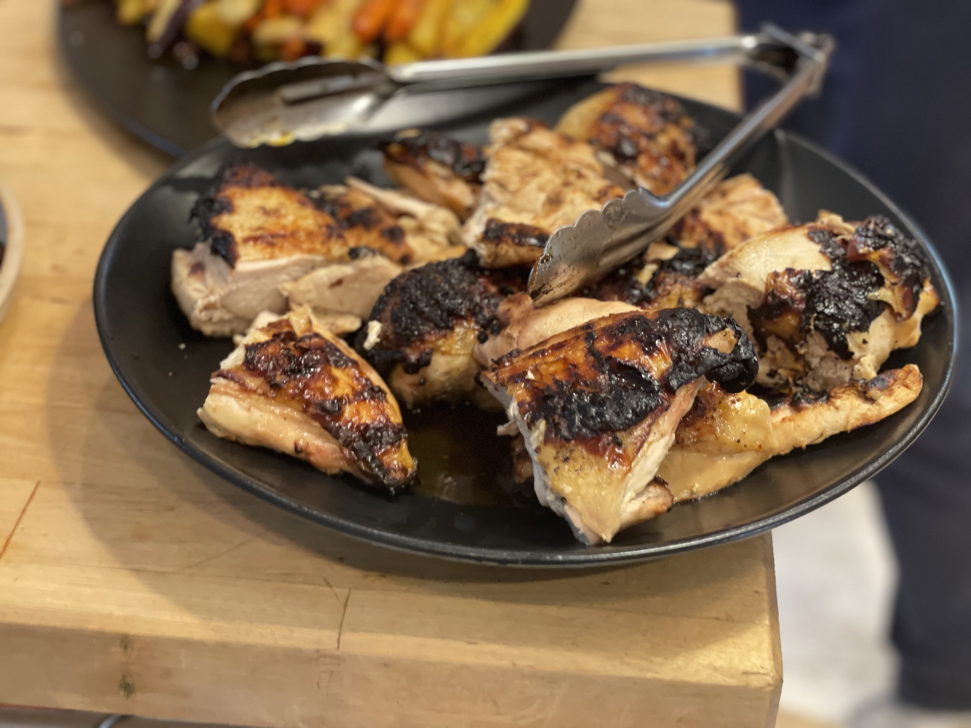 Charred grilled chicken pieces on a dark plate with tongs