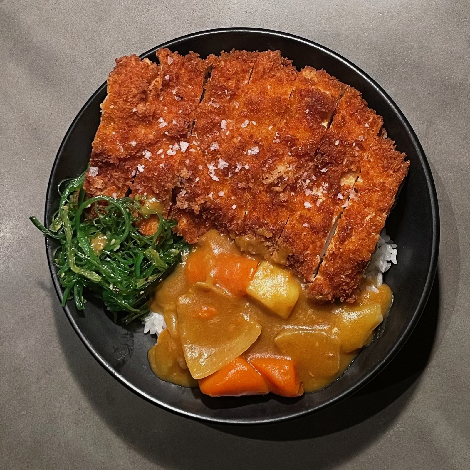Overhead view of katsu curry bowl with breaded cutlet, curry sauce, vegetables, and seaweed salad