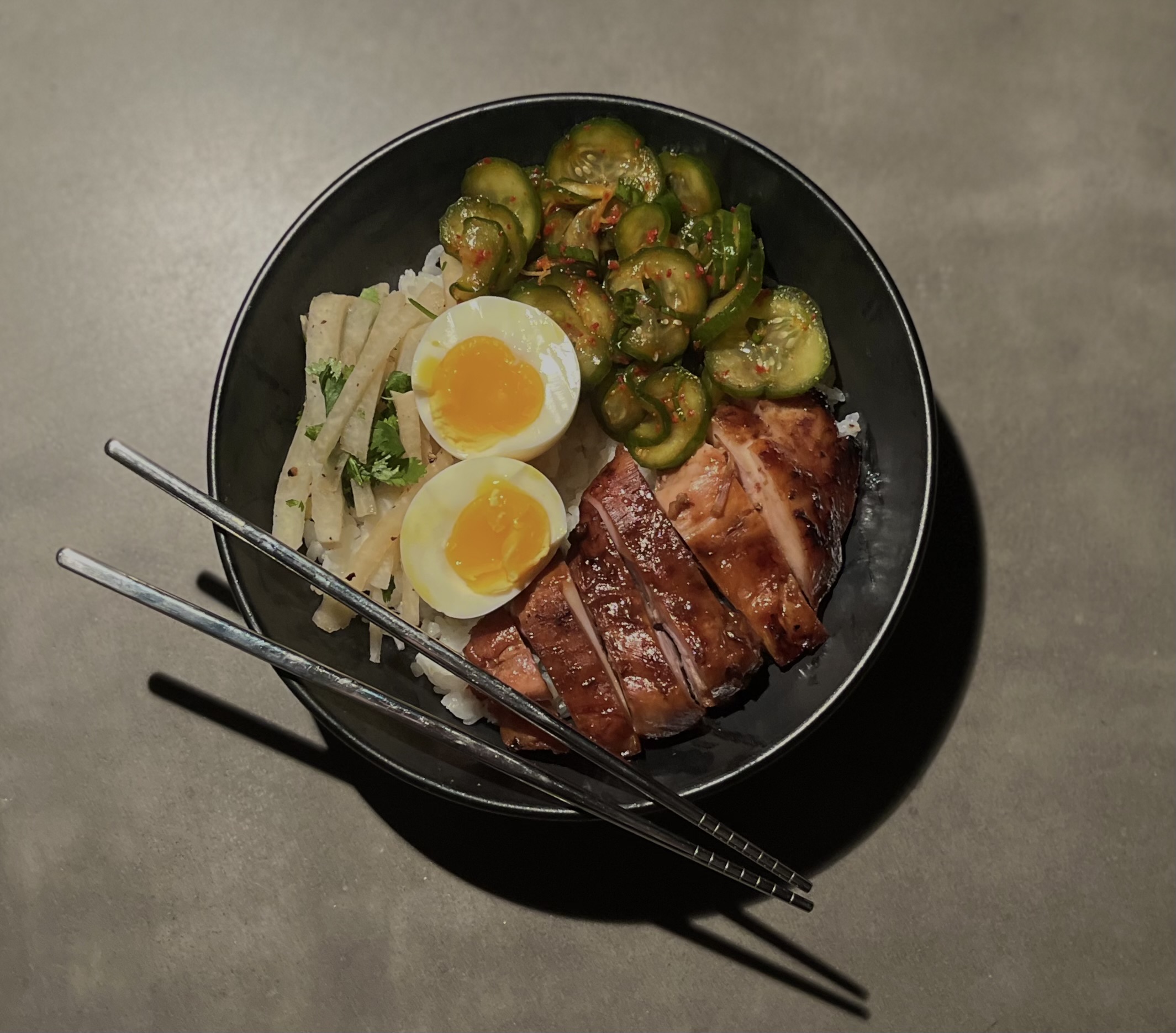 Rice bowl with glazed meat, soft-boiled egg, spicy cucumber salad, and pickled daikon