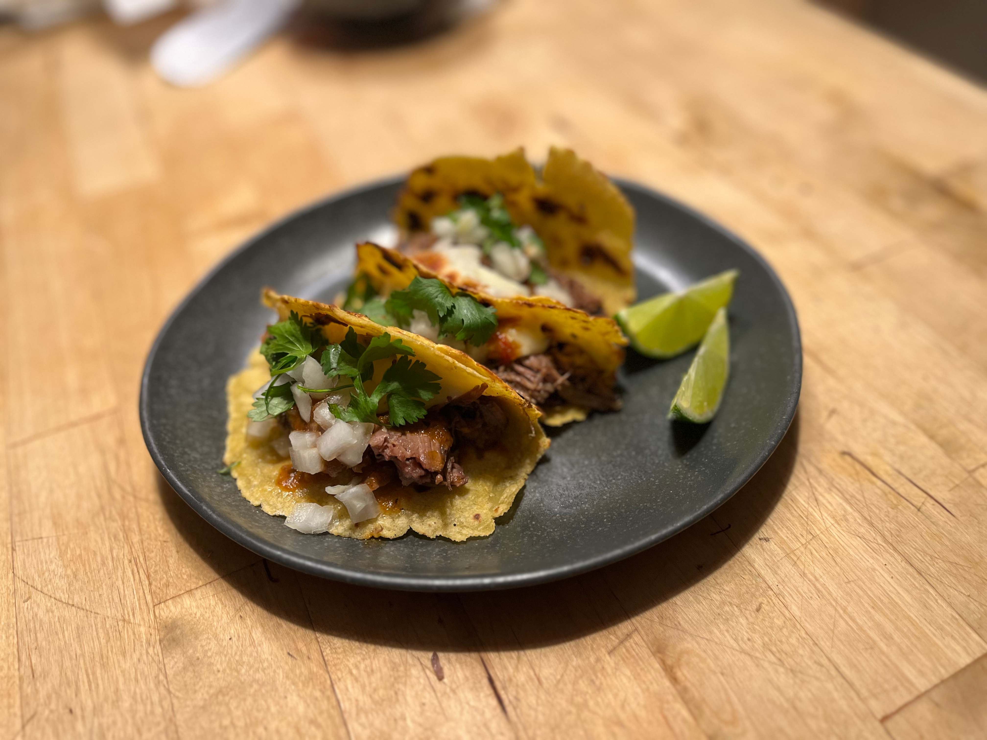 Corn tortilla tacos with braised meat, salsa, cilantro, onion, and lime on a dark plate